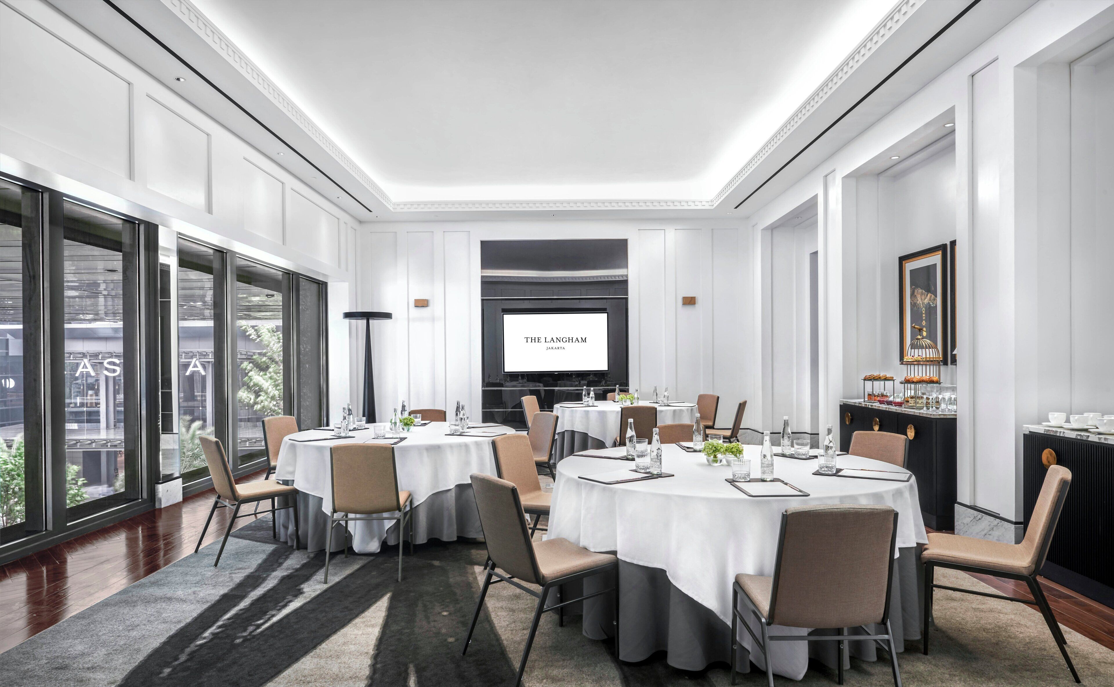 Una sala de conferencias con varias mesas, sillas y una pantalla proyectora mostrando 'THE LANGHAM JAKARTA'.