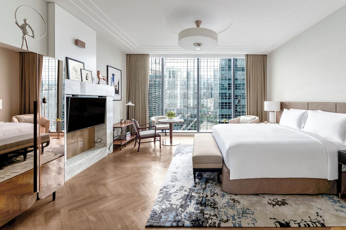 Quarto de hotel moderno com uma cama grande, televisão, escrivaninha com cadeira, mesa pequena com um vaso e vista da cidade através de janelas amplas.
