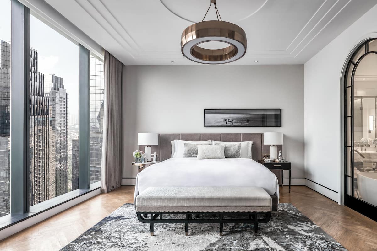 Quarto de hotel moderno com uma cama grande, vista para a cidade, um espelho amplo e uma luminária de teto circular.