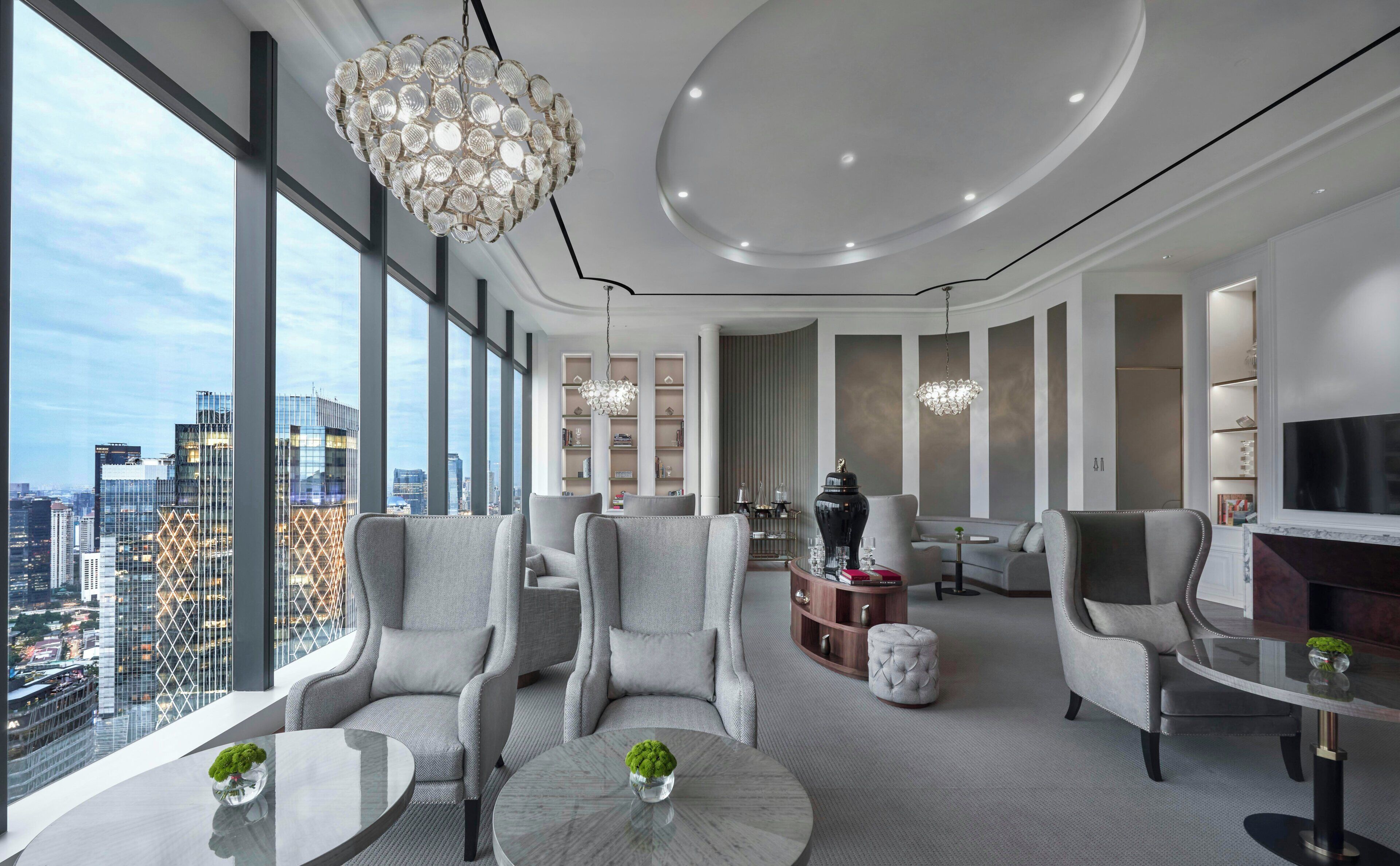 A modern living room with a city view, a round coffee table, grey upholstered chairs, a chandelier, and a fireplace.