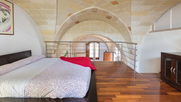 Ein Schlafzimmer mit Bett, Holzboden, roter Decke und Blick auf eine Küche durch einen Bogen.