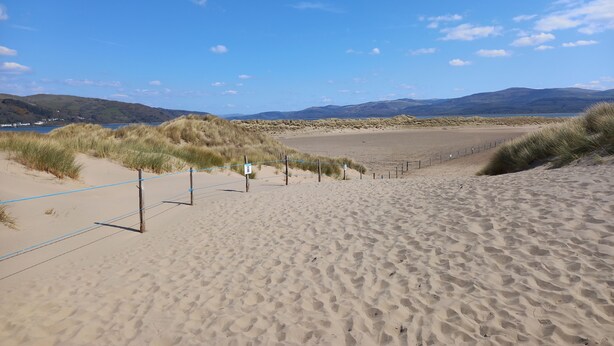 Una playa arenosa con cielo despejado, dunas cubiertas de hierba y montañas a lo lejos.