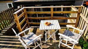 Una terraza de madera con una mesa y dos sillas, un plato pequeño con comida y una vista de un edificio al fondo.