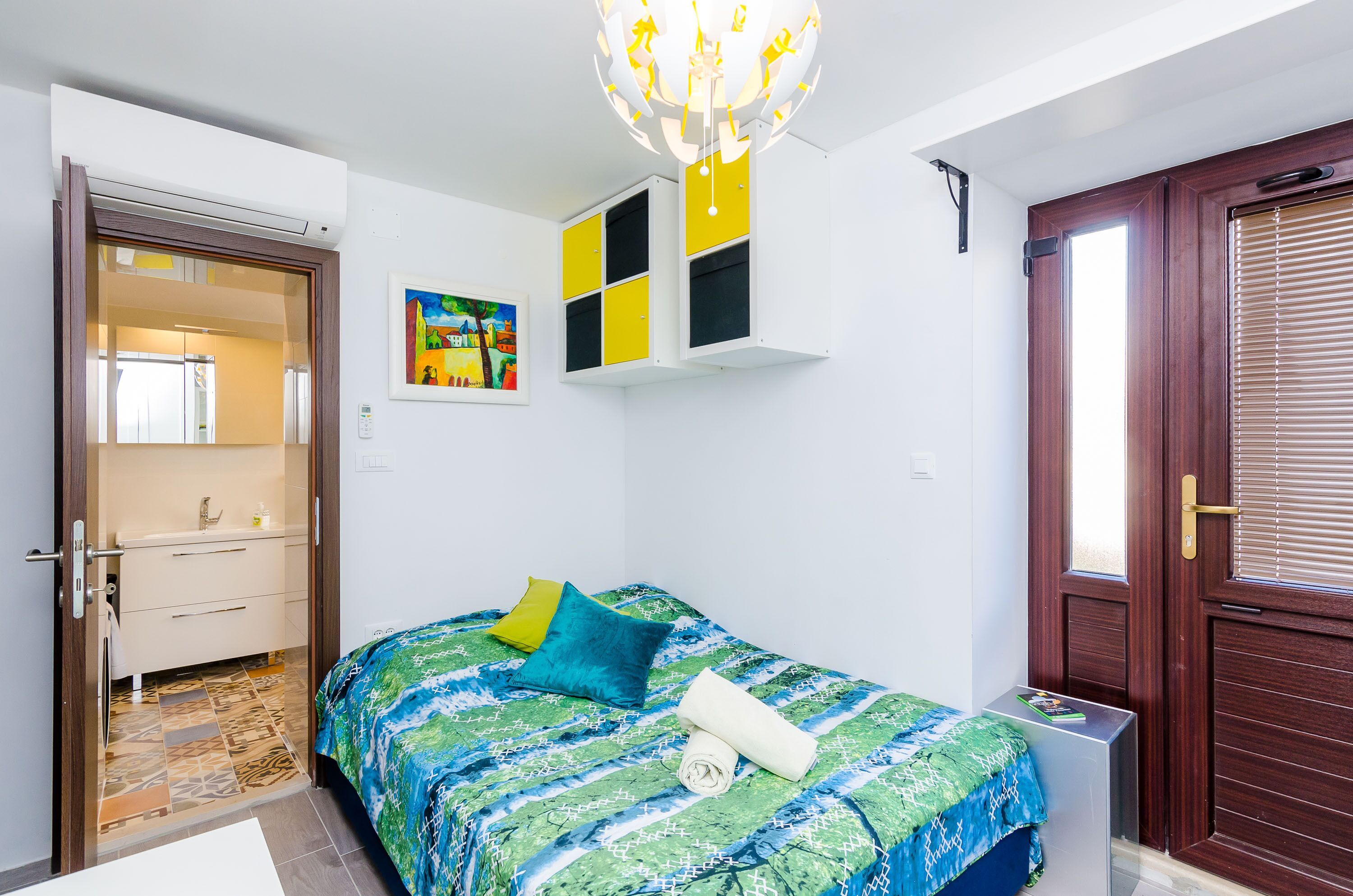 A bedroom with a bed, a door, a mirror, a ceiling light fixture, and a small table.