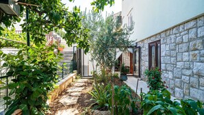 A narrow stone pathway flanked by lush greenery and potted plants, leading to a building with a stone facade and wooden doors. - Apartment & Room Sunset Beach (Dubrovnik)