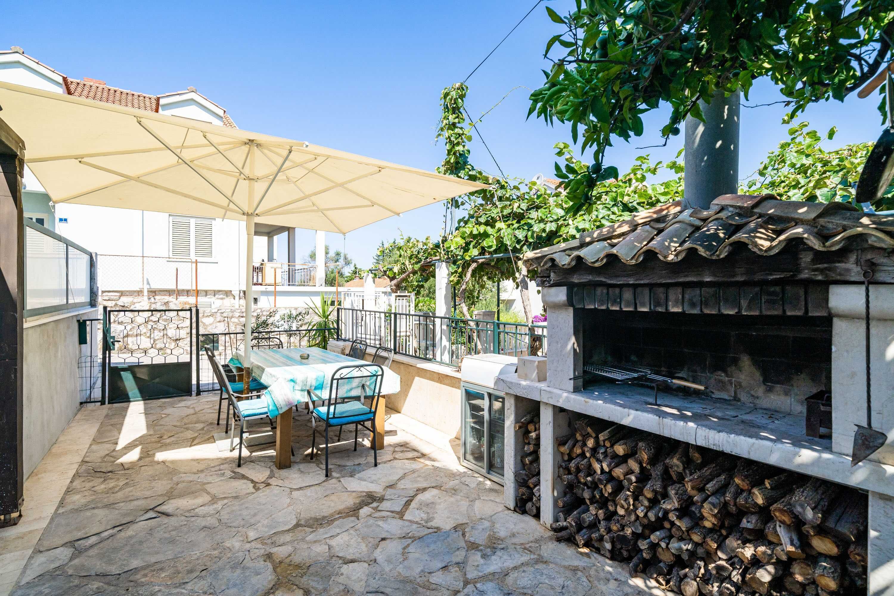 Une terrasse avec un sol en pierre, une table avec des chaises, un barbecue et un grand parasol.