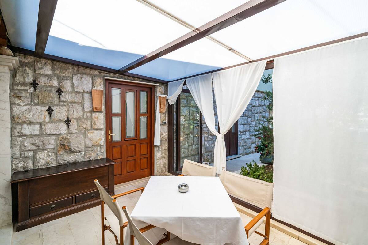 A covered patio with a stone wall, a wooden bench, a table with a white tablecloth, and a door with glass panels.