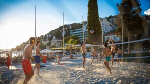 A beach volleyball game on a sandy court with a net, surrounded by beach umbrellas and people. - Apartment & Room Sunset Beach (Dubrovnik)