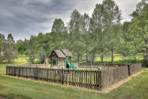 Une structure de jeu en bois avec un toboggan, entourée d’une clôture en bois, installée dans un espace verdoyant avec des arbres en arrière-plan.