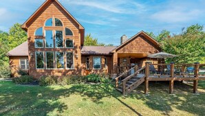 Une maison en bois à deux étages, dotée d’un grand porche à l’avant, d’une cheminée en pierre et d’une pelouse bien entretenue.