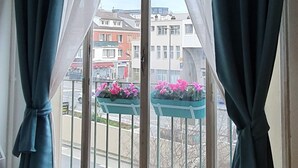 Una habitación con un ventanal grande, cortinas, una lámpara colgante y vistas a edificios y flores afuera.