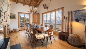 A dining area with a wooden table set for six, large windows with mountain views, stone walls, and rustic decor including antlers and skis.