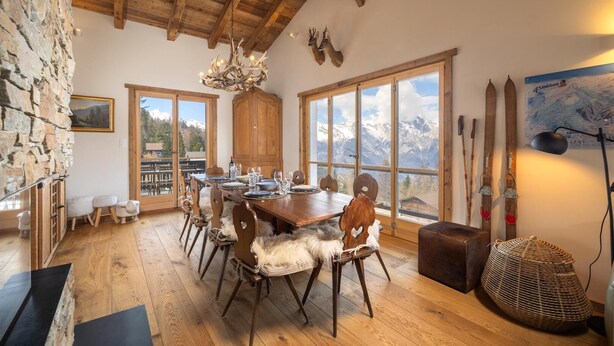 A dining area with a wooden table set for six, large windows with mountain views, stone walls, and rustic decor including antlers and skis. - Chalet Pain Seigle (Riddes)