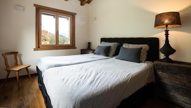 A bedroom with two beds, a wooden chair, a nightstand with a lamp, and a window with a view of trees.