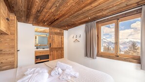 A bedroom with a large bed, wooden ceiling, a bathroom with a mirror, and a window with a mountain view. - Chalet Pain Seigle (Riddes)