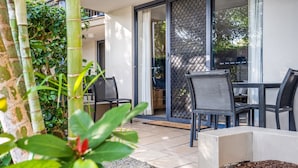 An outdoor patio with a table and chairs, a sliding glass door, and a bamboo plant. - 2-Bed Family Haven with Pools, BBQ & Playgrounds (Mermaid Beach)