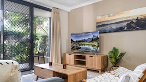 A living room with a TV, wooden furniture, a sofa, a chair, and a view of greenery outside. - 2-Bed Family Haven with Pools, BBQ & Playgrounds (Mermaid Beach)