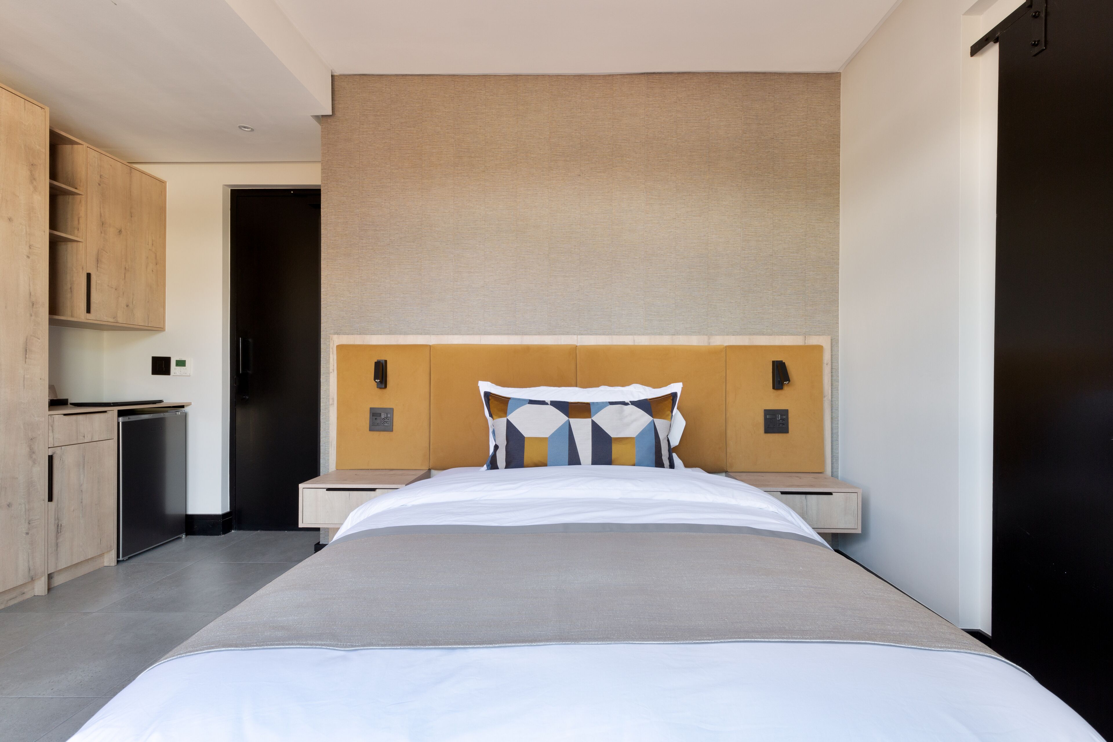 Una habitación de hotel moderna con una cama grande, un armario de madera, una pequeña kitchenette y una puerta oscura.