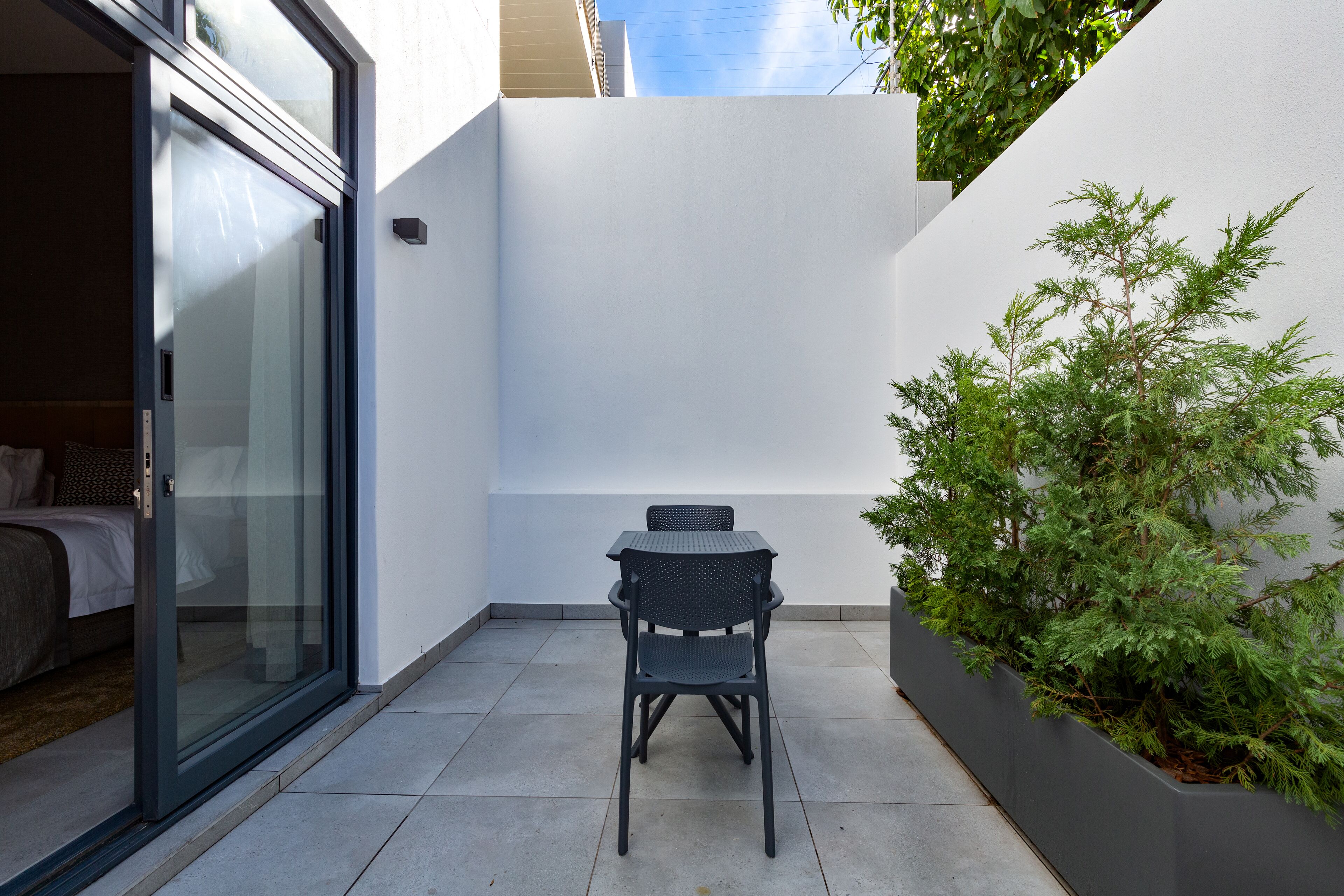 Un piccolo patio con tavolo e sedia, una porta vetrata scorrevole che dà su una camera da letto e un arbusto verde a contrasto con un muro bianco.