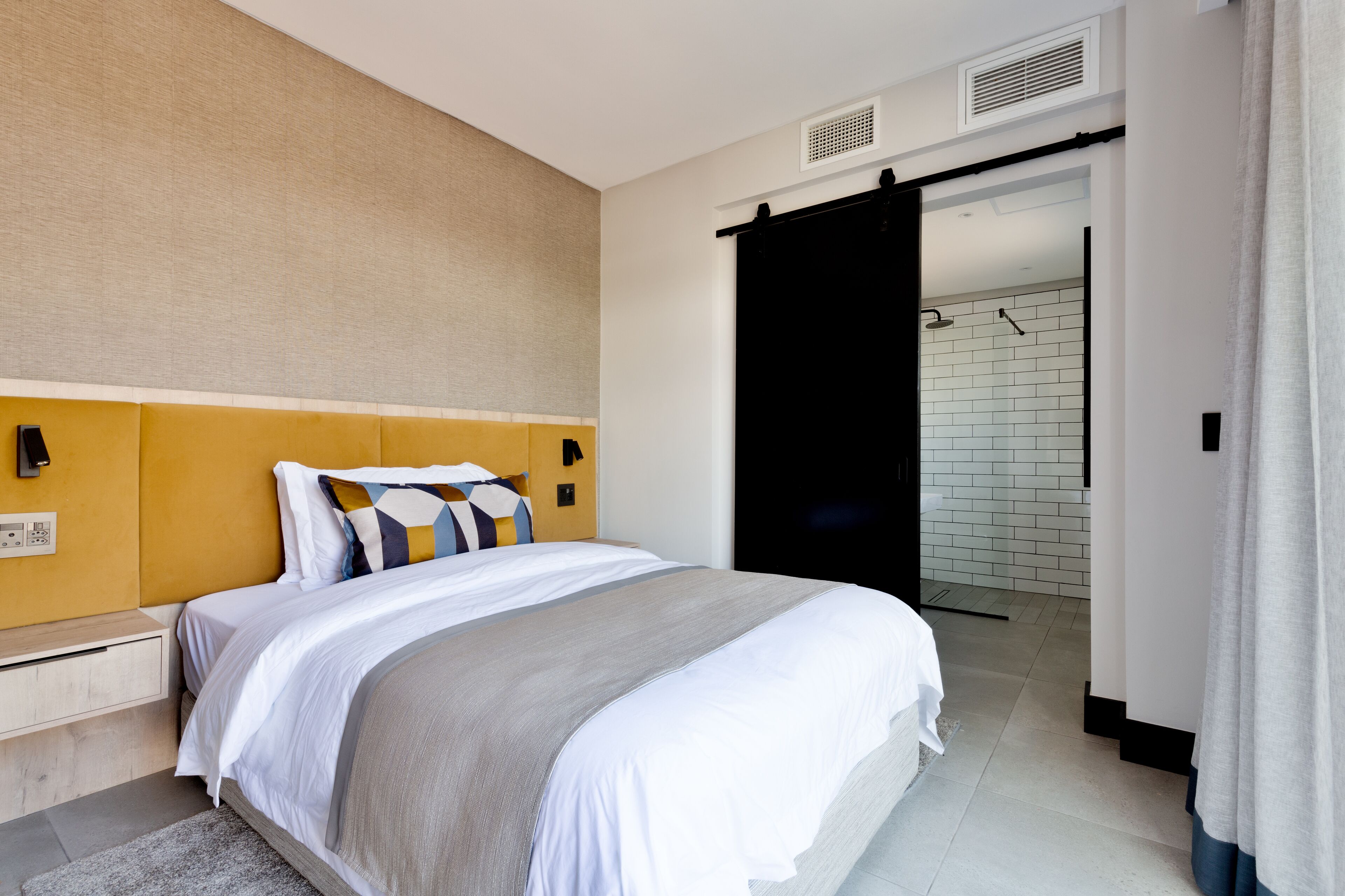 A modern hotel room with a large bed, a headboard, a bedside table, and a bathroom visible through an open door.