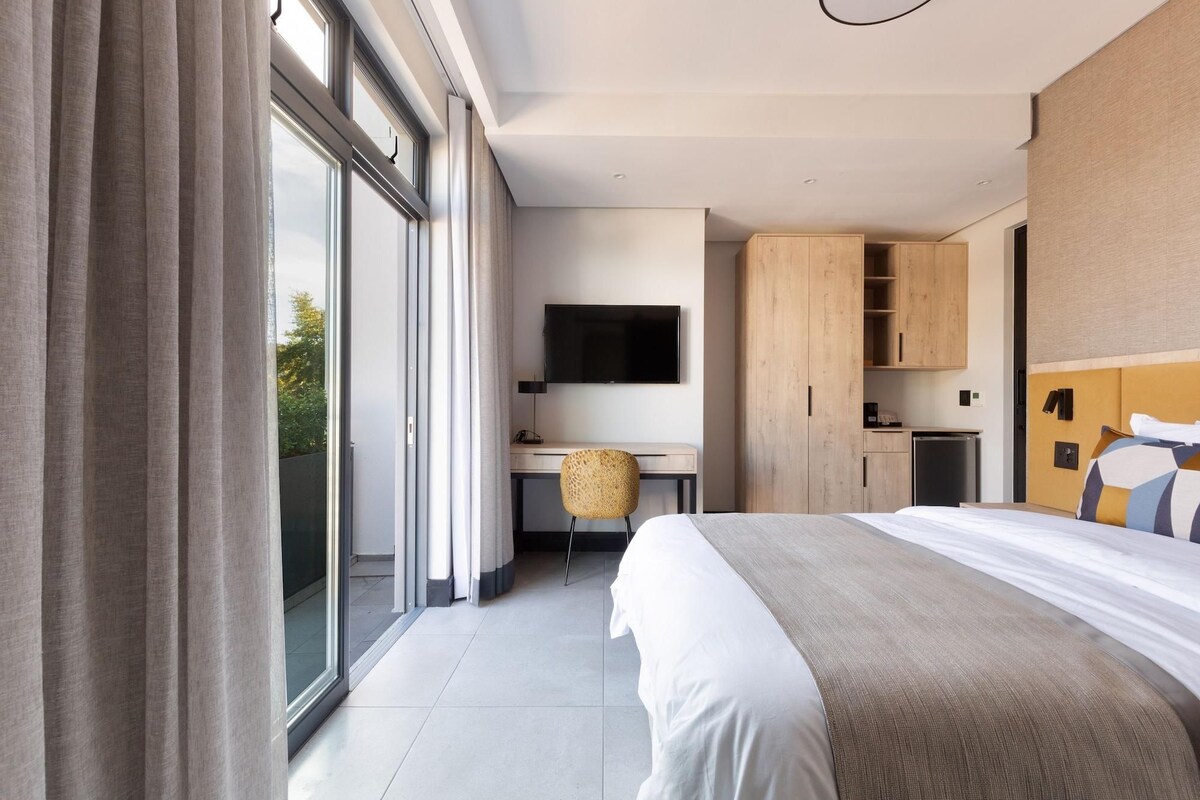 A modern hotel room with a large bed, a desk with a chair, a television, and a kitchenette.