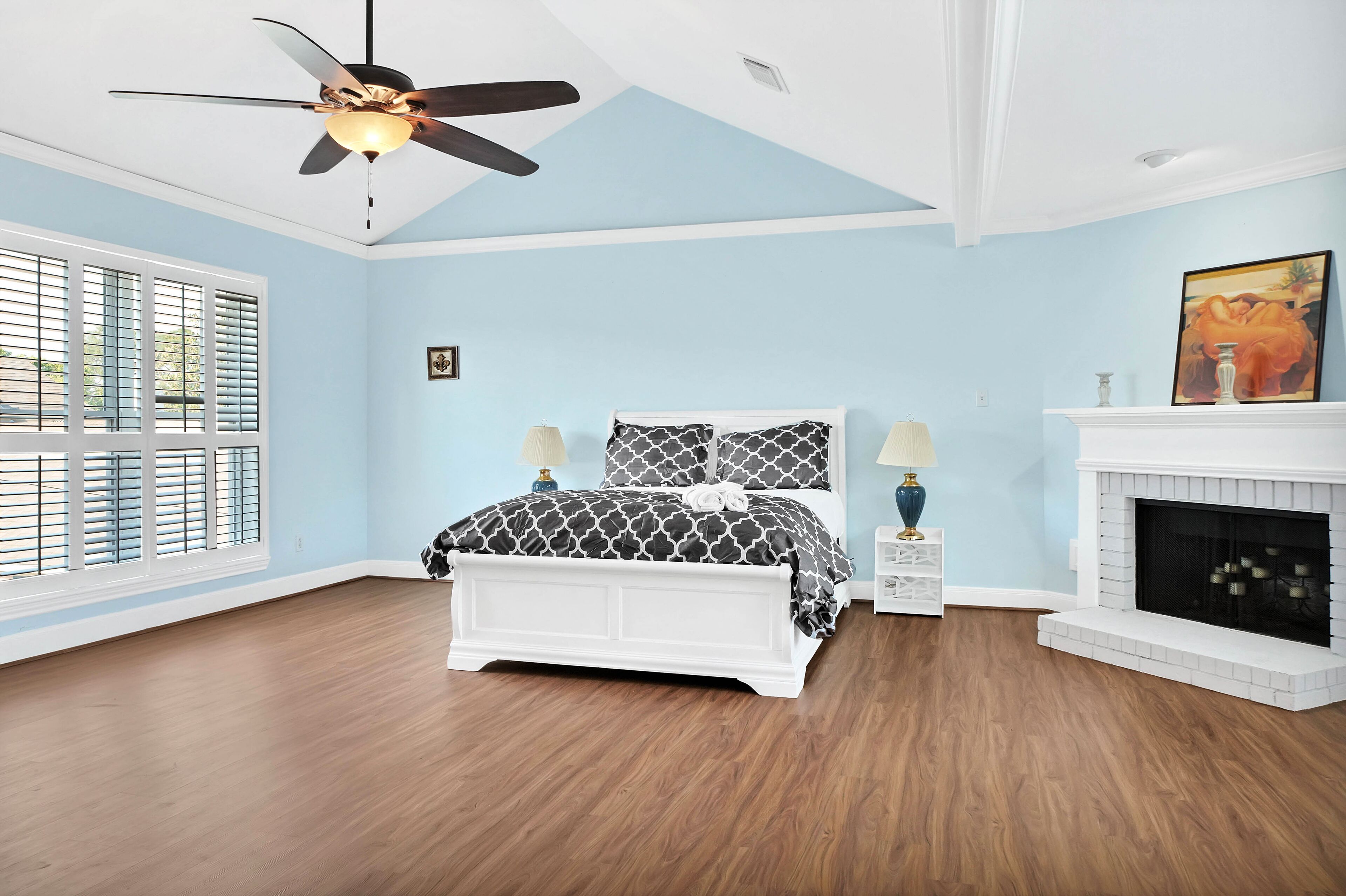 A bedroom with a bed, two bedside tables, a ceiling fan, a fireplace, and a large window with blinds.