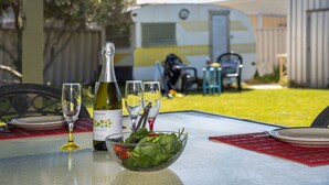 A table with a bottle of sparkling wine, glasses, and a bowl of salad in an outdoor setting with a trailer in the background.