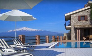 Un espace aménagé au bord de la piscine, avec des chaises longues et un parasol, un bâtiment en pierre doté d’un balcon, et une montagne en arrière-plan.