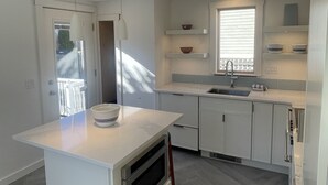 Una cocina moderna con armarios blancos, una isla central, iluminación colgante y un ventanal con persianas.