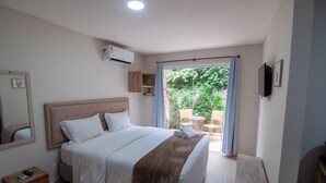 Un dormitorio con cama, mesita de noche, un pequeño cofre de madera, televisor y ventana con cortinas.