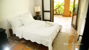A bedroom with a bed, a nightstand, a lamp, and a door leading to an outdoor area with plants.
