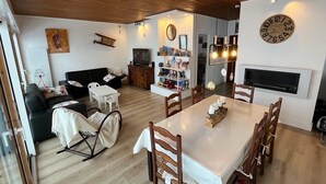 A cozy living room with a sofa, a dining table, and a TV. There is a bookshelf and a wooden ceiling.