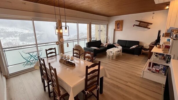 A spacious living area with a wooden ceiling, a dining table with chairs, a sofa, and a small white table.