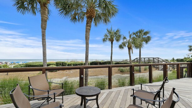 A wooden deck with four chairs and a small table, overlooking a beach with palm trees and a clear blue sky. - Beautiful renovated home in the heart of Sea Pines with Private Pool! (Hilton Head Island)