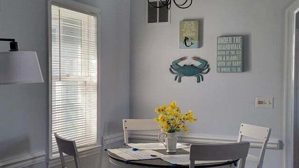 A dining area with a round table, white chairs, a vase with yellow flowers, and wall decorations including a crab and a book. - 25b Olive Ave.- North Rehoboth Cottage with prime ocean-block location (Rehoboth Beach)