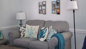 A grey sofa with patterned cushions, a floor lamp, and two framed pictures on the wall. - 25b Olive Ave.- North Rehoboth Cottage with prime ocean-block location (Rehoboth Beach)