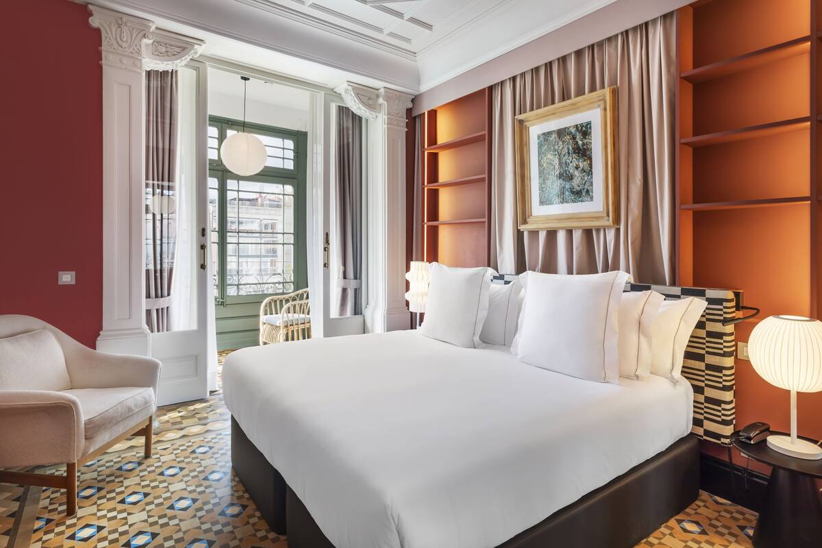 A hotel room with a large bed, a chair, a nightstand with a lamp, and a view of the outside through a window.