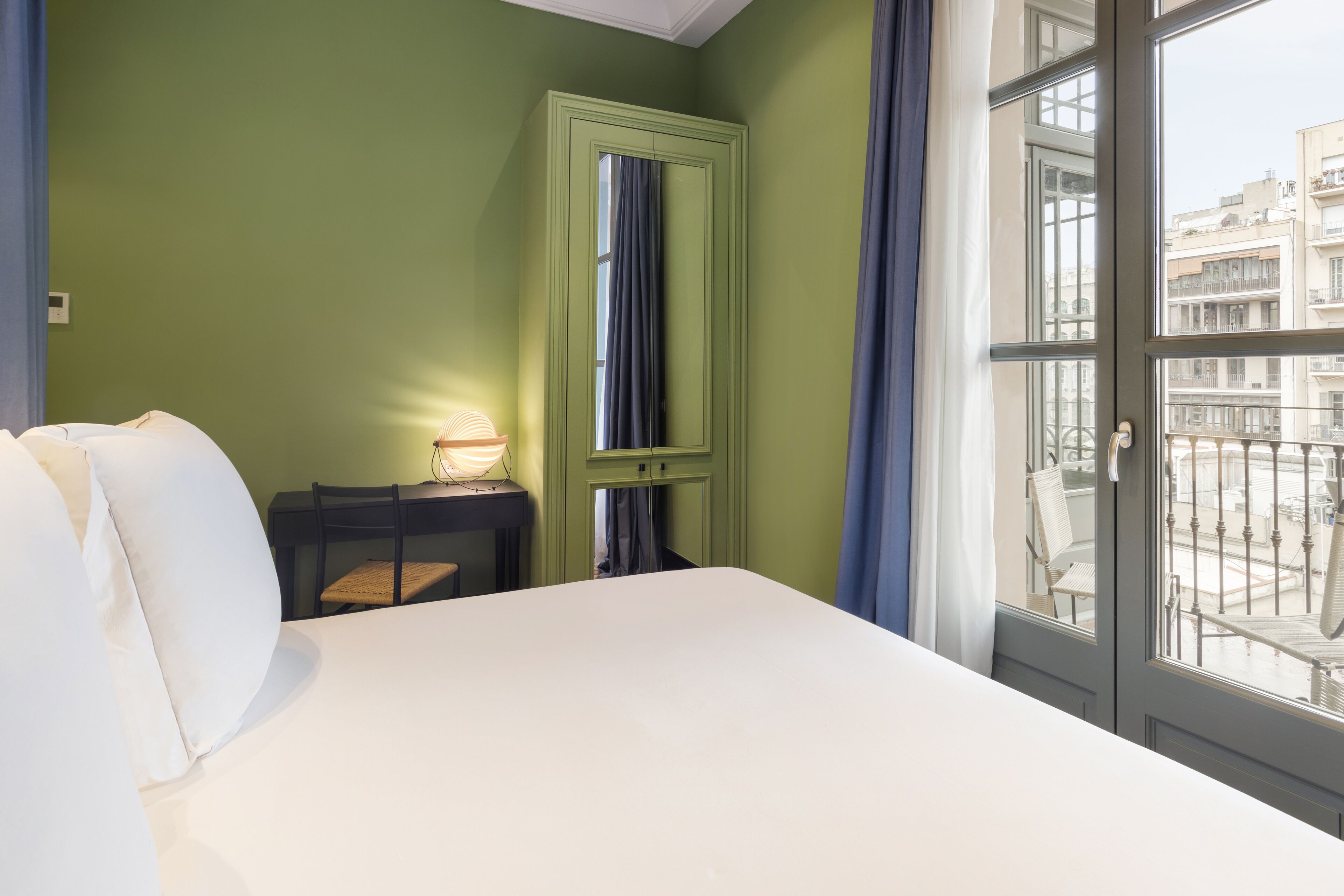 A hotel room with a large bed, a desk with a lamp, a mirror, and a view of the city through a window.
