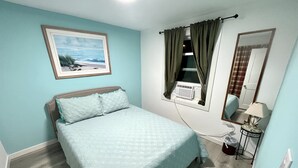 Un dormitorio con cama, ventana con cortinas, mesita de noche con lámpara, espejo y un cuadro en la pared.