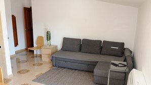 A modern bedroom with a grey sofa bed, a small wooden side table, a chair, and a TV stand with a remote. - Cetara House 1 on Amalfi Coast (Cetara)