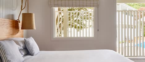 Un dormitorio con cama, lámpara colgante, ventana con persianas y vista a una cerca blanca y follaje exterior.