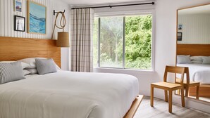 A bedroom with a large bed, a wooden chair, a window with a view of greenery, and a mirror reflecting the bed. -  Parker Beach Lodge (South Yarmouth)