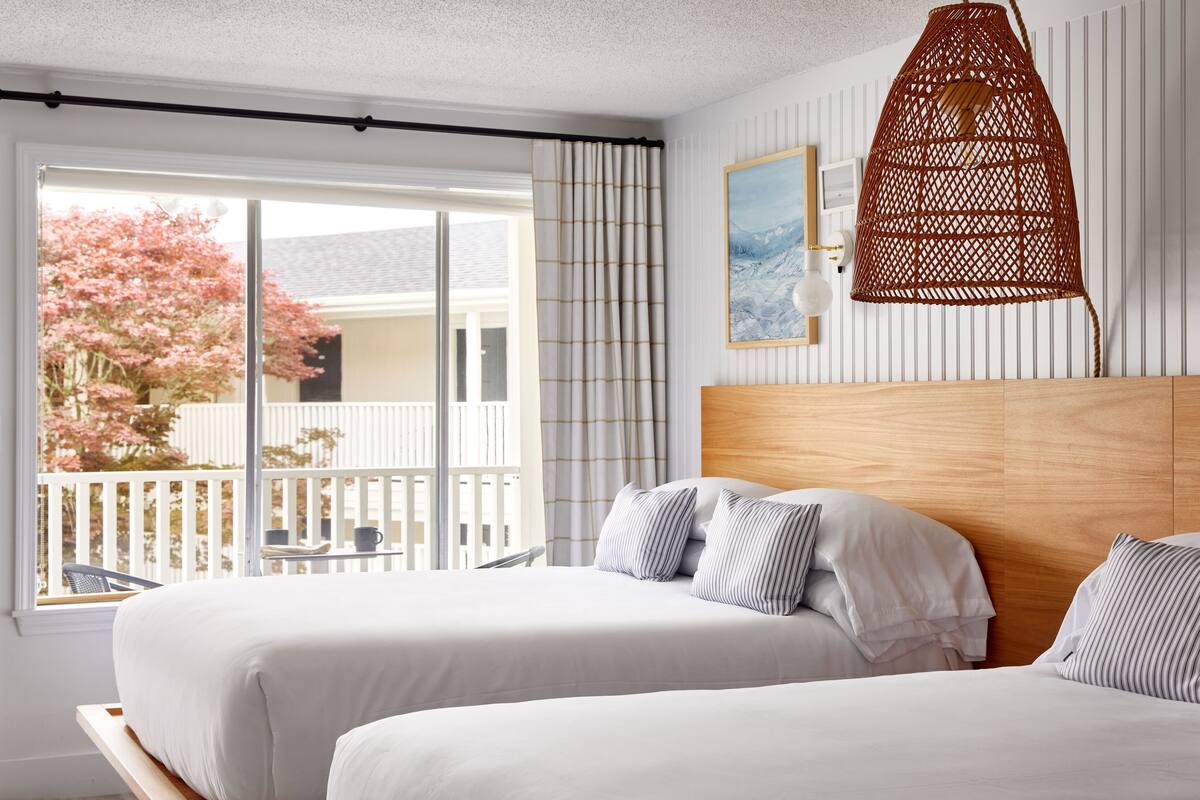 Hotellihuone, jossa on kaksi sänkyä, puinen pääty, suuri ikkuna, josta on näkymä parvekkeelle ja vaaleanpunaisiin puihin, sekä roikkuva, punottu valaisin.