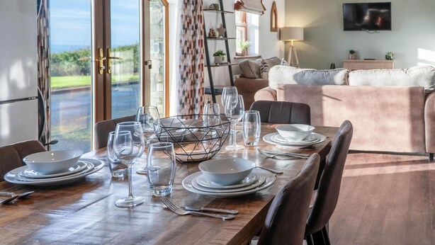 A dining area with a wooden table set for four, a sofa in the background, and a view through the glass doors.