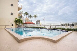 A swimming pool with a white fence, palm trees, and a building in the background. - The Blue Octopus - 1br King Bed Condo (Fort Lauderdale)