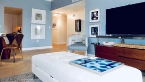 Una sala de estar moderna con un televisor de pantalla plana grande, un sofá blanco y una mesa de centro con un diseño abstracto azul. Hay cuadros enmarcados en las paredes y un aparador de madera con objetos decorativos.