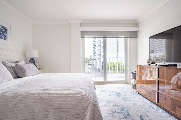 A hotel room with a bed, a television, a dresser, and a balcony with a view of buildings.