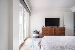A bedroom with a bed, a television on a wooden dresser, a window with blinds, and a trash bin.