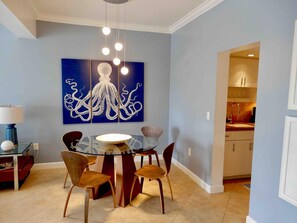 Un coin repas avec une table en verre entourée de chaises en bois, un tableau bleu représentant une pieuvre accroché au mur, et une cuisine visible à travers une porte ouverte.