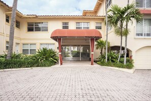 A building entrance with a covered waiting area, palm trees, and a gated driveway.